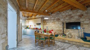 A rustic interior with stone walls, wooden beams, a dining area with green cushioned chairs, and a television mounted on the wall. - AKAMANTIDA PLATEA 2. Country house of the 1900s. Restored to its authentic charm (Pano Arodes)