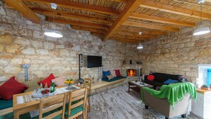 A rustic stone-walled room with a wooden ceiling, a fireplace, a sofa, and a dining area with a table and chairs. - AKAMANTIDA PLATEA 2. Country house of the 1900s. Restored to its authentic charm (Pano Arodes)