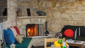 A stone-walled room with a fireplace, a sofa with colorful cushions, a wooden table with a vase of flowers, and a television on a stand. - AKAMANTIDA PLATEA 2. Country house of the 1900s. Restored to its authentic charm (Pano Arodes)