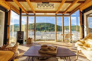 A cozy room with a wooden ceiling, large windows, a fireplace, and a view of a hill. - Quinta Maresia Tiny House 2 (Carvoeira)