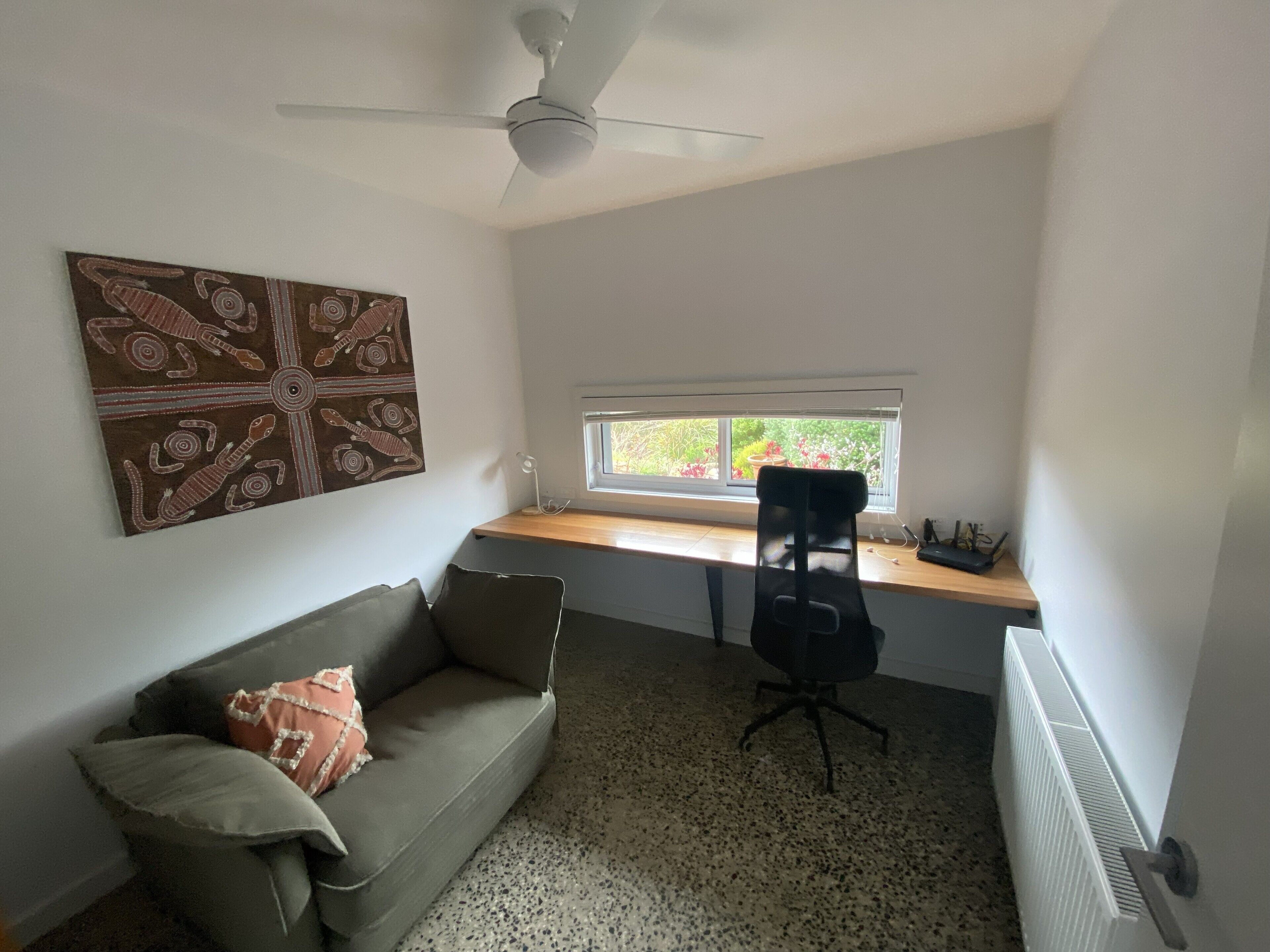 A small, bright room with a ceiling fan, a window with a view of greenery, a desk with a chair, and a couch with pillows.