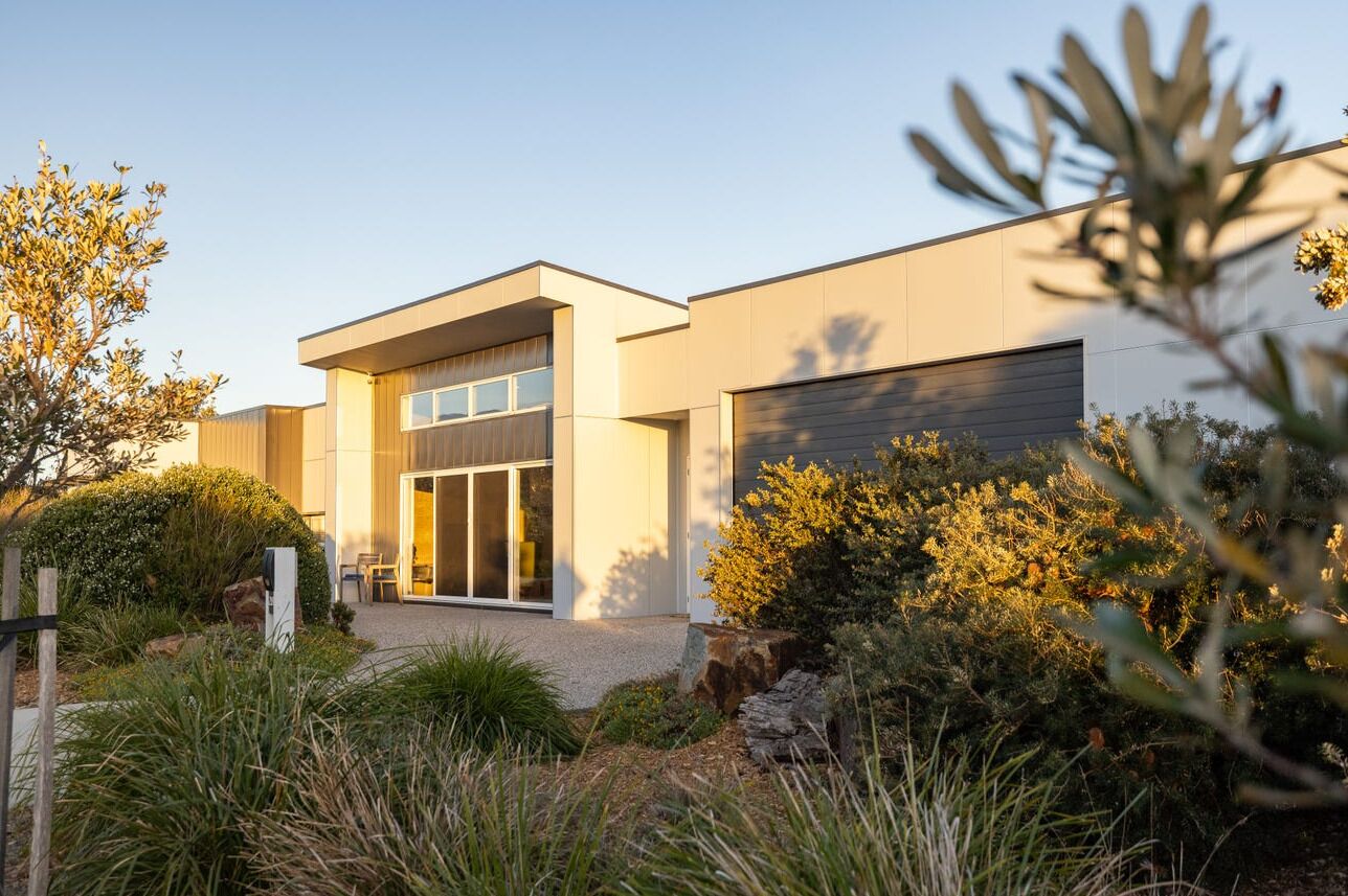 A modern two-story house with a large garage door, surrounded by a landscaped garden with various plants and shrubs.
