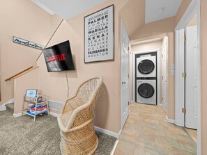 Un pasillo con un televisor montado en la pared, un cartel en la pared, una silla de mimbre, un área de lavandería con dos lavadoras y una puerta.