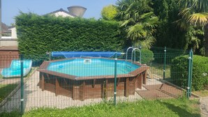 Een zwembad omgeven door een houten terras en een metalen hek, met een achtergrond van weelderige begroeiing en een grote kegelvormige constructie.