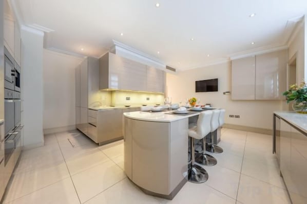 A modern kitchen with a central island, built-in appliances, and a flat-screen TV mounted on the wall. - Royal Mayfair Villa (London)