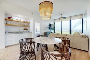 A modern kitchen with white cabinets, a dining area with a round table and wicker chairs, a living room with a beige sofa, and large windows offering a view. - High-End 2BR, Resort Perks: Suntide III - Unit 107 (South Padre Island)