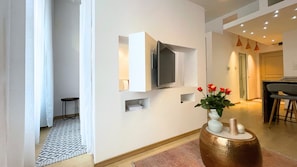 Uma sala de estar moderna com uma televisão de tela plana na parede, um vaso com flores vermelhas sobre uma mesa lateral de madeira e um tapete estampado no chão.