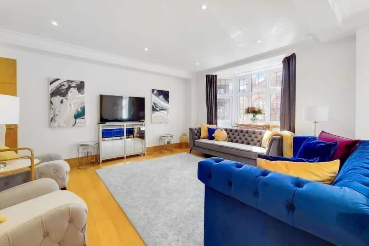 A modern living room with a blue sofa, a grey sofa, a flat-screen TV, artwork on the walls, and a window with curtains.