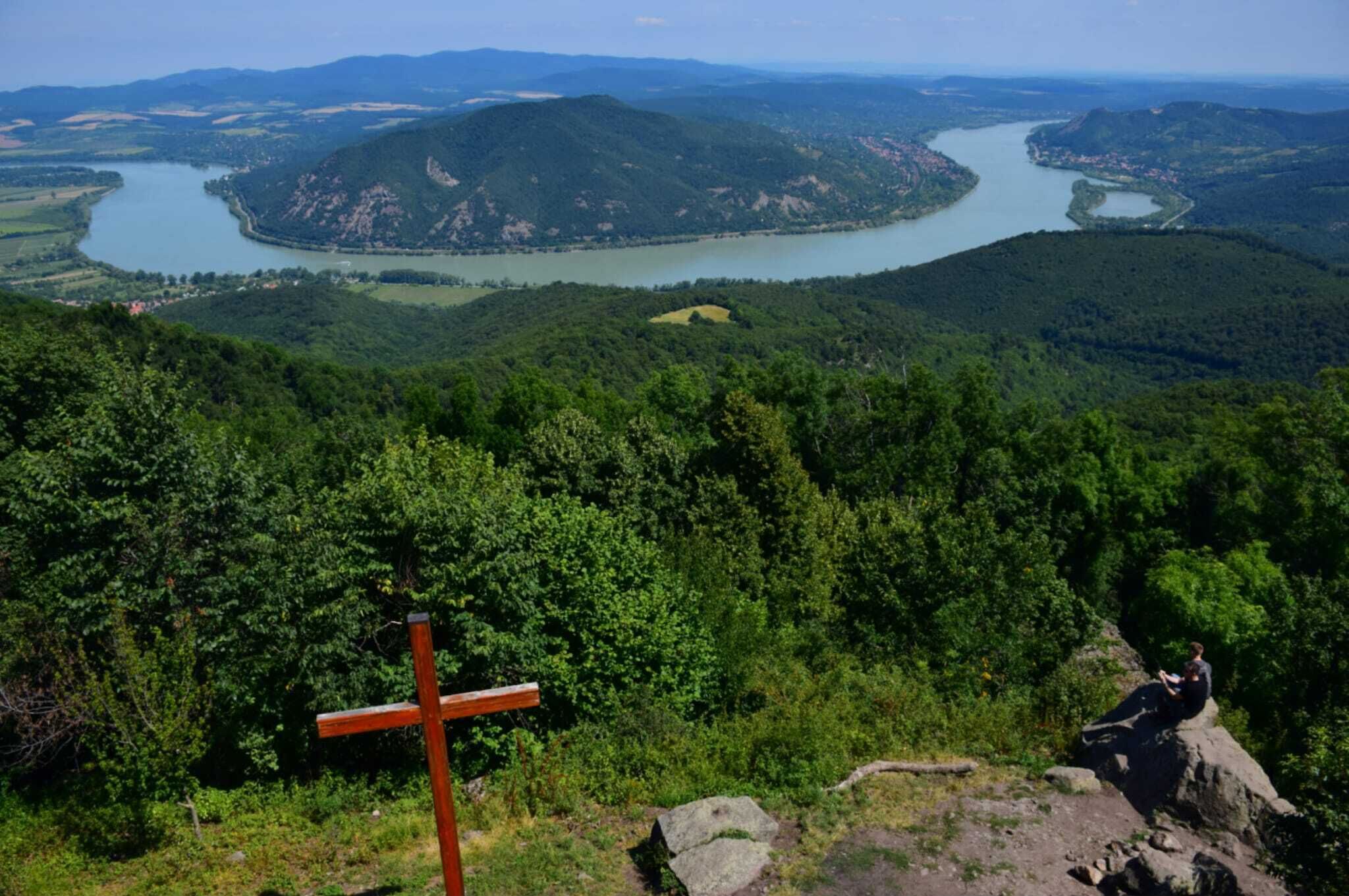 A cross in a forested area with a view of a river and mountains in the distance.