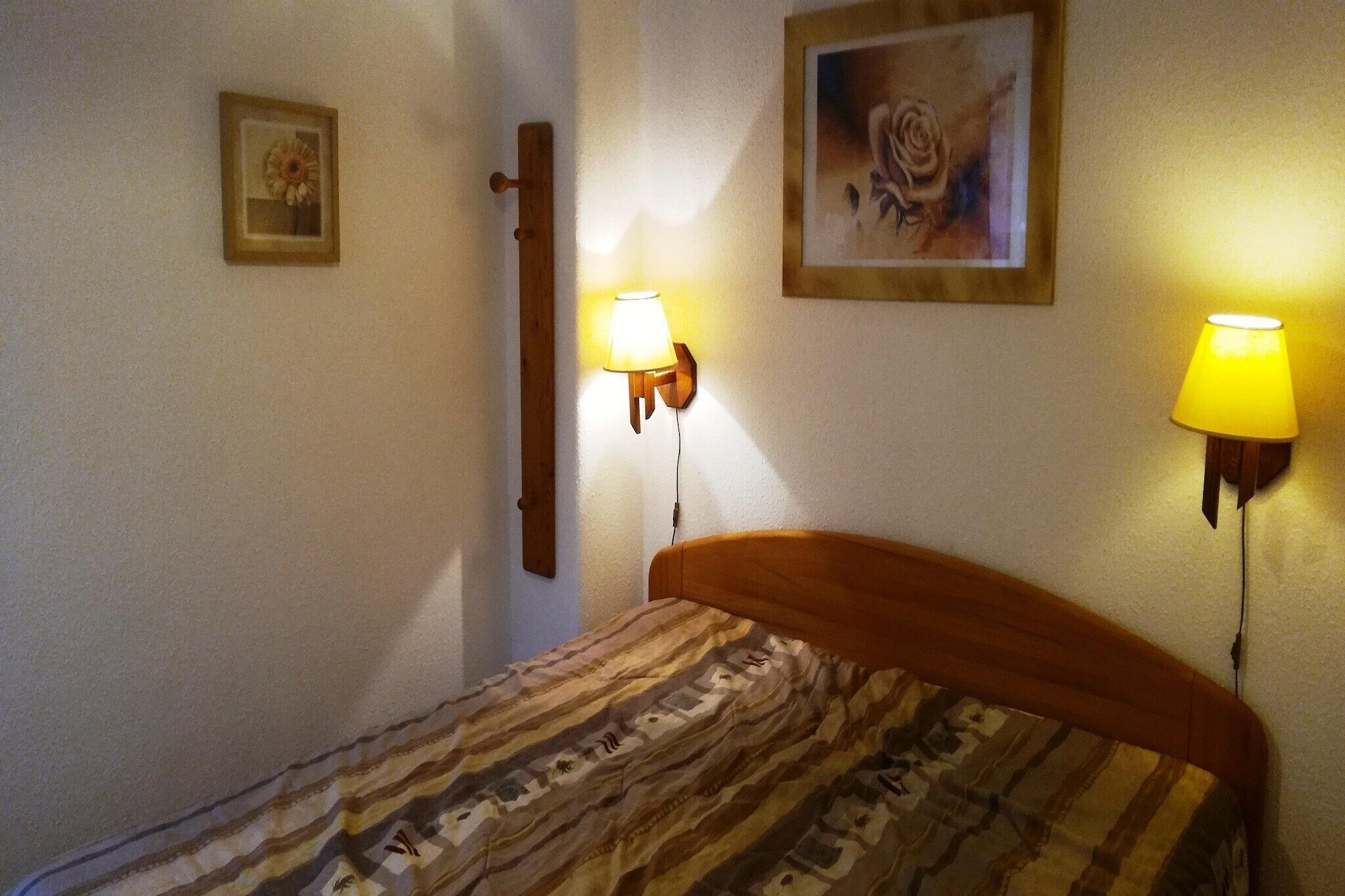 A bedroom with a wooden headboard, a patterned bedspread, two wall-mounted lamps, and two framed pictures on the wall.