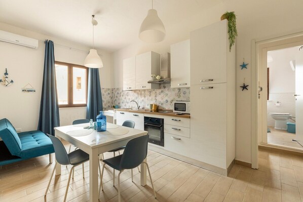 A modern kitchen with white cabinets, a dining table with chairs, a blue sofa, and a hanging plant. - Appartamento con aria condizionata (Sassari)