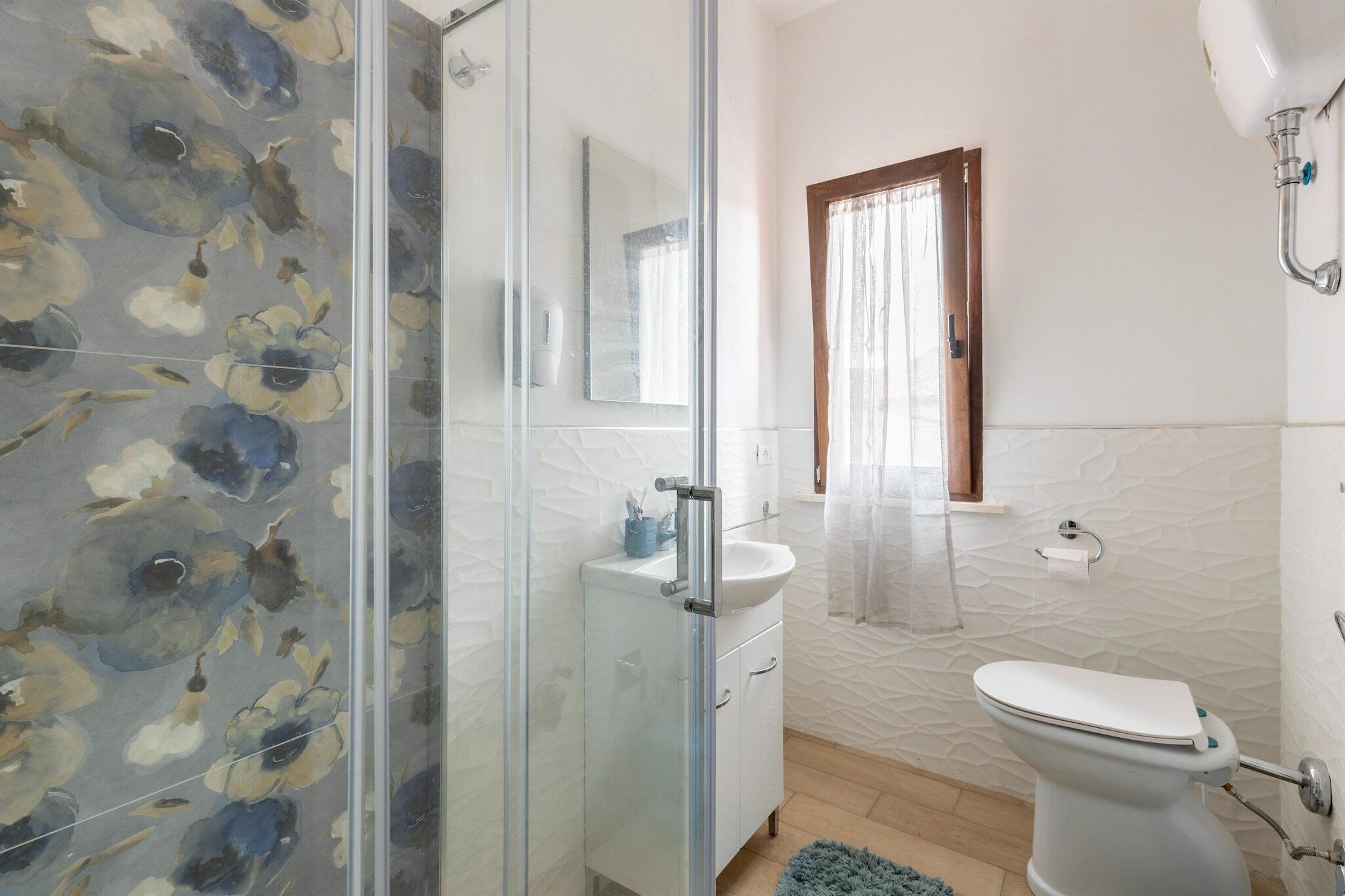 Un baño con cabina de ducha de vidrio, lavamanos blanco, inodoro y ventana con marco de madera.