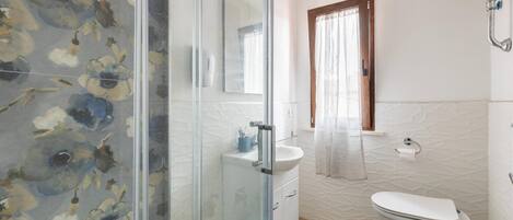Un baño con cabina de ducha de vidrio, lavamanos blanco, inodoro y ventana con marco de madera.