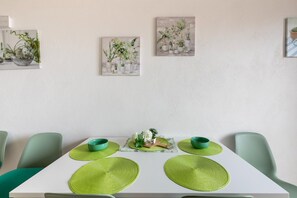 Ruokailutilassa on valkoinen pöytä, vihreät aluslautasliinat ja tuolit. Seinällä on kolme taulua, joissa on kuvattuna kasveja.