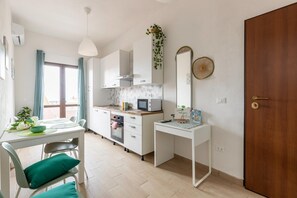 Eine moderne Küche mit weißen Schränken, einer Mikrowelle und einem Spülbecken. Ein Esstisch mit Stühlen und ein kleiner Schreibtisch mit Spiegel sind ebenfalls vorhanden.