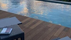 Una piscina infinita con cubierta de madera y sillones de descanso, con vistas a un atardecer sobre una masa de agua.