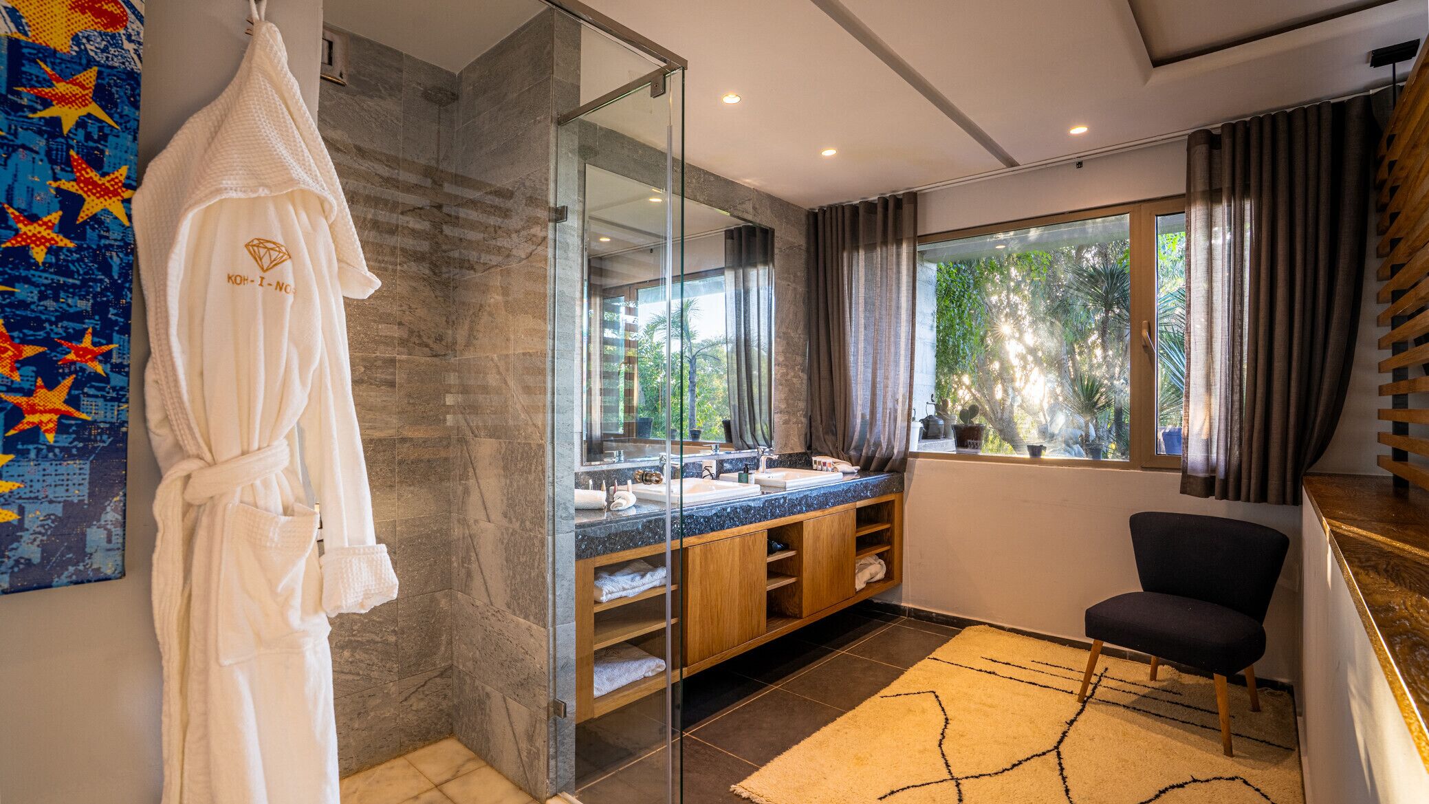 Un baño moderno con cabina de ducha de vidrio, tocador doble y un amplio ventanal con vistas a la vegetación.