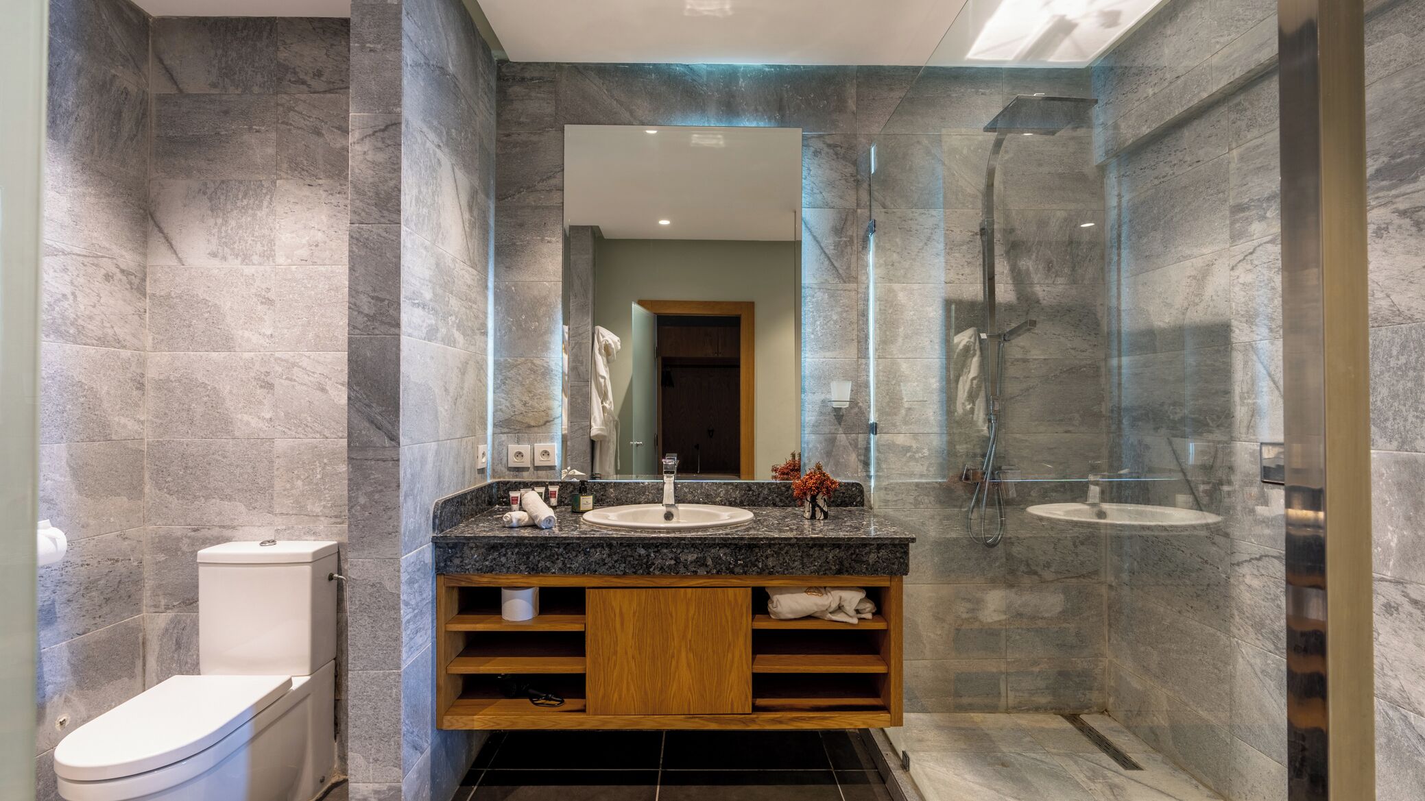 Un baño moderno con encimera de granito, tocador de madera, un espejo grande y ducha con mampara de vidrio.