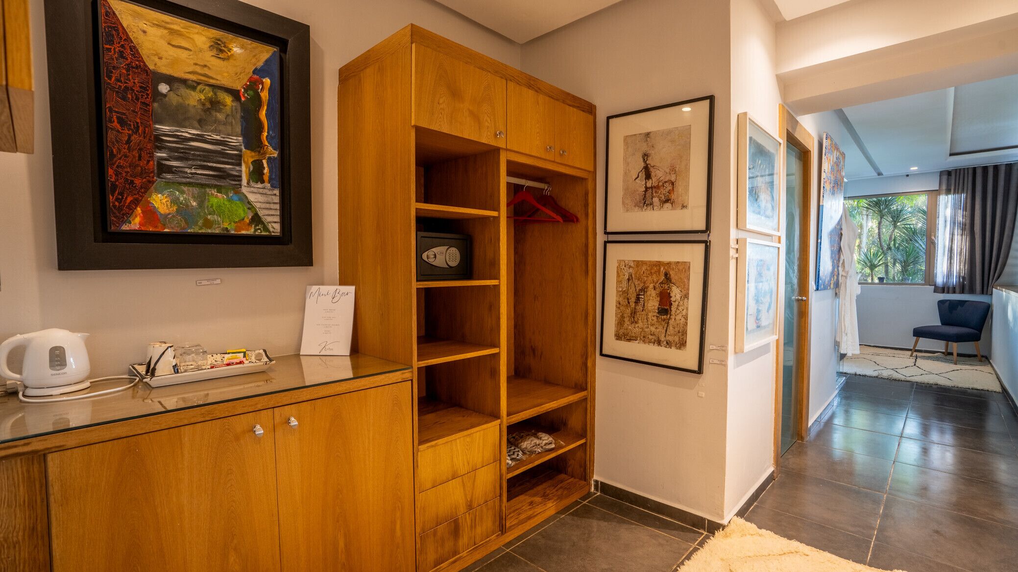A hotel room with a wooden cabinet, a framed abstract painting, a minibar, and a view of the outdoors.