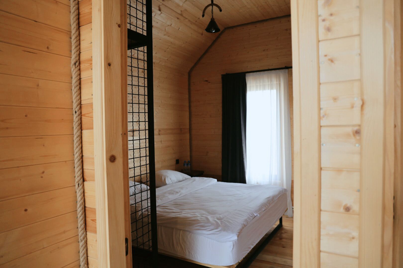 A wooden cabin room with a bed, a rope, and a window with curtains.