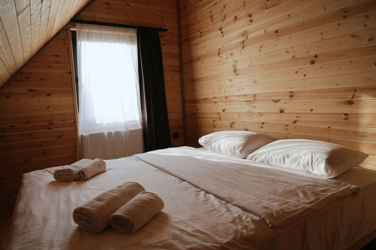Un dormitorio de cabaña de madera con una cama, dos almohadas y una ventana con cortinas.