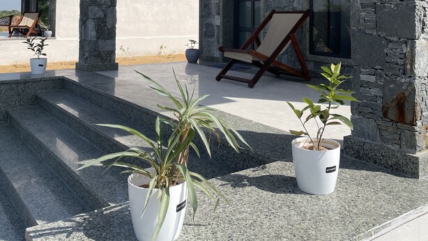 Un bâtiment moderne aux murs en pierre et au toit plat. Des marches mènent à un patio couvert où se trouvent une chaise et des plantes en pot.