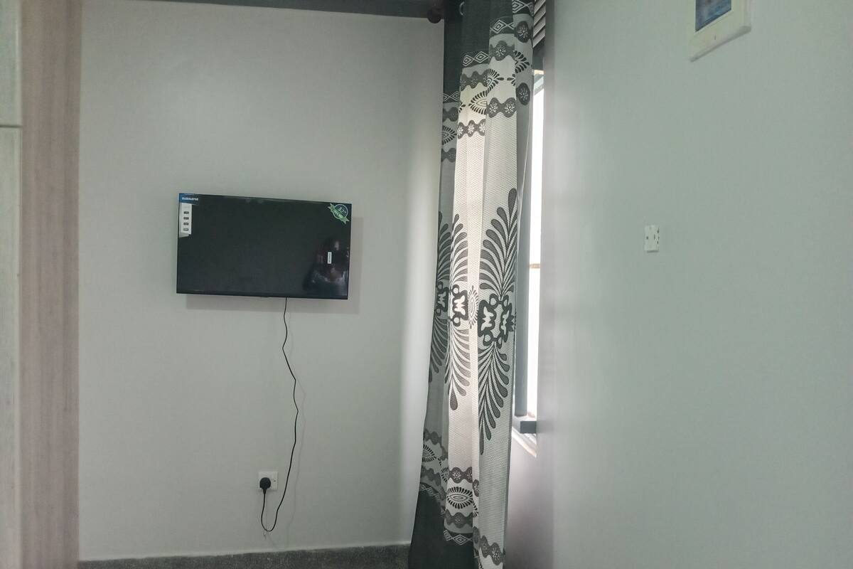 Habitación con televisor de pantalla plana montado en la pared, un enchufe y una ventana con cortina estampada.