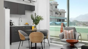 A modern kitchen with dark cabinets, a dining area with two chairs, a small table, and a view of a tennis court through large windows. - דירת סטודיו עם מרפסת במתחם סי סייד -By Edomlux (Eilat)