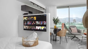 A modern hotel room with a large wall-mounted TV displaying multiple streaming channels, a wicker baby crib with a white blanket, a dining area with a small table and chairs, and a view of the mountains through large windows. - דירת סטודיו עם מרפסת במתחם סי סייד -By Edomlux (Eilat)