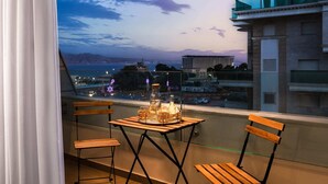 A balcony with a table and chairs, overlooking a cityscape and a body of water during twilight. - By Edomlux סטודיו מודרנית במתחם סי סייד 100 מטר מהים (Eilat)