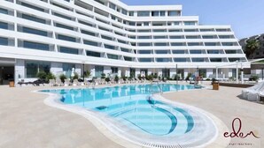 Ett modernt hotell med ett stort poolområde, flera balkonger och en klar himmel.