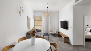 A modern living room with a dining table, chairs, a sofa, a TV, and a window with curtains. - דירה משפחתית מפנקת 100 מטר מהים - By Edomlux (Eilat)