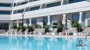 Ein moderner Hotelpoolbereich mit mehreren Liegestühlen und Sonnenschirmen, umgeben von einem mehrstöckigen Gebäude mit großen Fenstern.