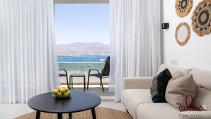Ein Zimmer mit Balkon, das auf ein Gewässer und Berge blickt. Es gibt einen runden Tisch mit Obstschale, eine Couch mit Kissen und eine Wand mit gewebter Dekoration.