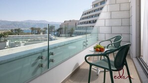 Un balcón con baranda de vidrio, dos sillas verdes, una mesita con fruta y vistas a un paisaje urbano y montañas al fondo.