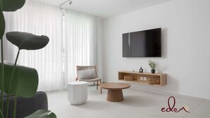 Una sala de estar moderna con un ventanal grande, un televisor de pantalla plana, una mesa de centro redonda, una mesita auxiliar blanca, una silla de madera y un aparador de madera con cestas.