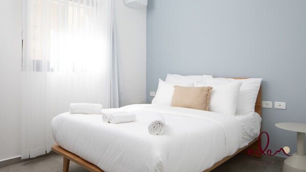 A neatly made bed with white linens and a beige throw pillow, a white bedside table, and a window with sheer curtains allowing natural light.