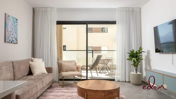 Uma sala de estar moderna com sofá, poltrona e mesa de centro, uma varanda com área de jantar e uma televisão de tela plana na parede.
