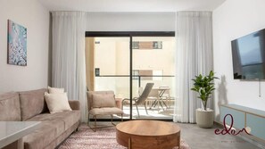 A modern living room with a sofa, armchair, and coffee table, a balcony with a dining area, and a flat-screen TV mounted on the wall.