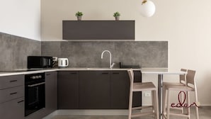Cozinha moderna com armários escuros, bancada branca, forno embutido, micro-ondas e máquina de café. Há duas cadeiras e uma mesa pequena.