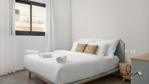 Cama bem arrumada com roupa de cama branca e toalhas enroladas, mesa de cabeceira com abajur e janela com cortinas.
