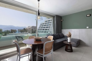 Un salon moderne comprenant un coin repas, un canapé, une petite table avec un vase, et une vue sur un immeuble et des montagnes.