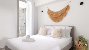 Quarto com cama, roupa de cama branca, toalha enrolada, peça de macramé na parede e uma pequena planta sobre um suporte de madeira.