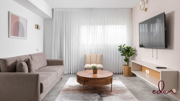 A modern living room with a sofa, a round coffee table, a TV, and a large window with sheer curtains. - דירה משפחתית בסי סייד בריכה מחוממת - By Edomlux (Eilat)
