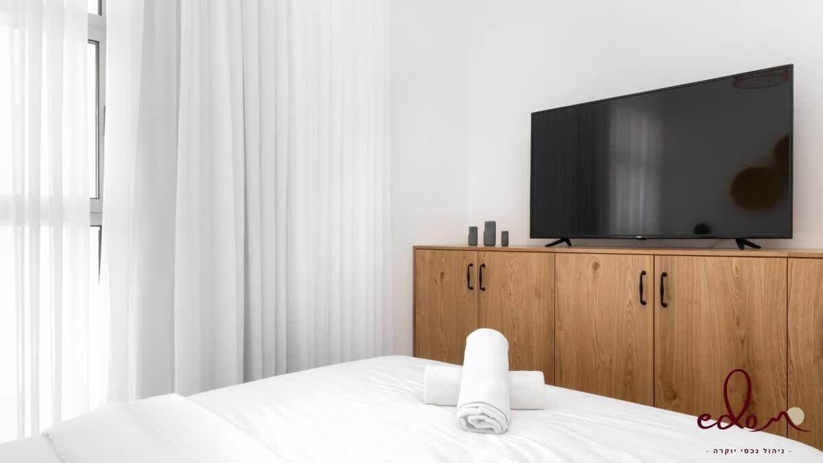 A hotel room with a bed, a wooden wardrobe, a flat-screen TV, and a window with sheer curtains.