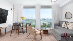 A modern living room with a sofa, chairs, a coffee table, and a TV. There are large windows with curtains, a view of a body of water, and a cityscape in the background. - דירה בסי סייד עם נוף מדהים לים - By Edom (Eilat)