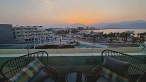 A balcony with two wicker chairs and a small table, overlooking a cityscape with buildings and a body of water during sunset. - דירה בסי סייד עם נוף מדהים לים - By Edom (Eilat)