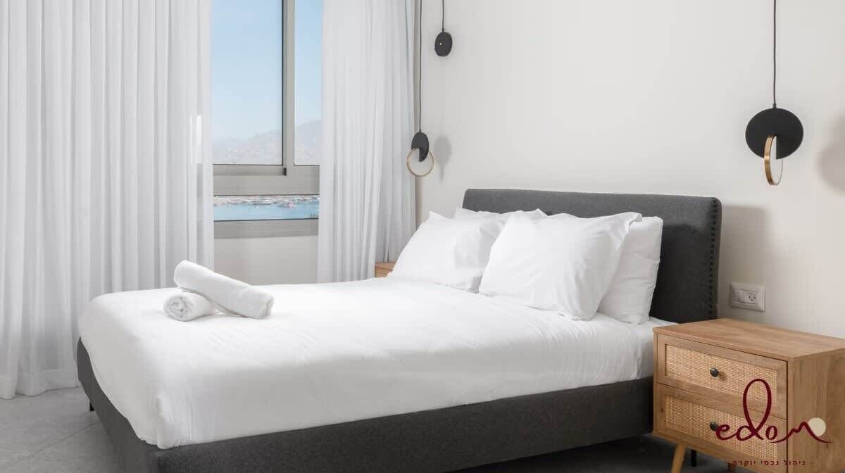 A neatly made bed with white linens and a rolled towel, a bedside table with a woven design, and a window with curtains.