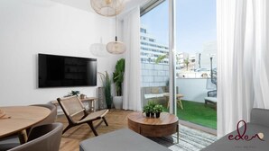 Een moderne woonkamer met een flatscreen televisie, rieten meubels, een ronde salontafel en een uitzicht op een stadslandschap door grote ramen.