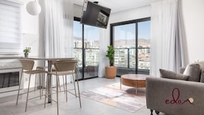 Uma sala de estar moderna com um sofá, uma mesa de jantar com cadeiras, uma TV fixada na parede e uma janela grande com vista para a cidade.